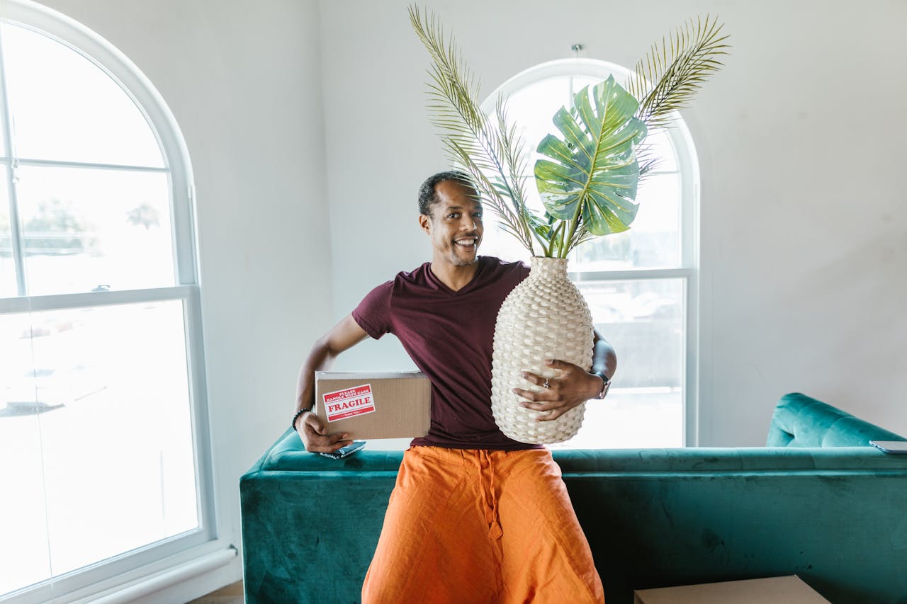 Smiling man holding a fragile box and plant during a home move.